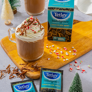 Box of Tetley Indulgence Chocolate & Mint Tea standing next to an elaborate latte with whipped cream topping, candy cane pieces, and chocolate flakes. 