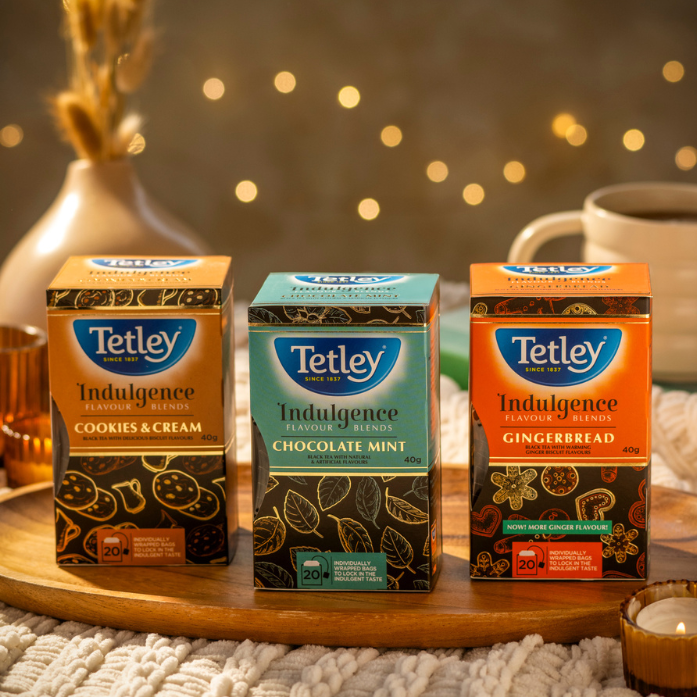 Tetley Indulgence Teas displayed on a wooden board surrounded by cozy accents like candles, and a soft blanket. 