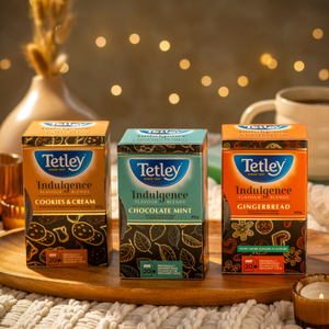 Tetley Indulgence Teas displayed on a wooden board surrounded by cozy accents like candles, and a soft blanket. 