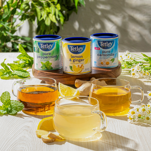 Tetley Pure Peppermint, Lemon & Ginger, and Pure Camomile tea canisters displayed on a wooden board and surrounded by fresh ingredient cues and cups of steeped tea. 